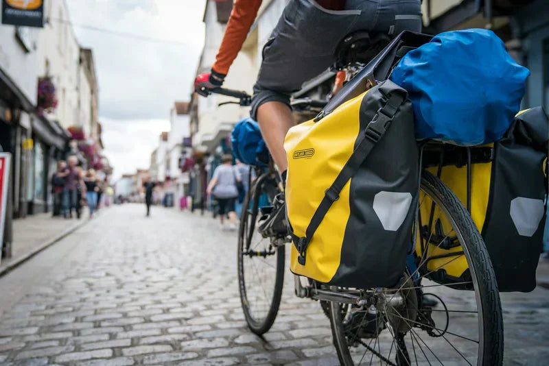 A cyclist with large yellow and blue panniers on their bike, riding on a cobbled street.