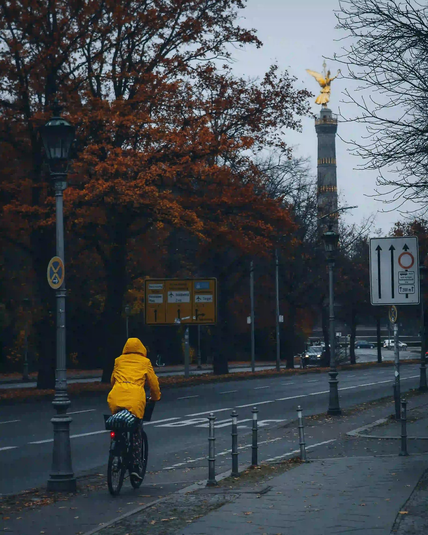 A person in a yellow raincoat rides an e-bike through Berlin in autumn, with the Victory Column in the background, illustrating the need for all-weather e-bike insurance in Germany.