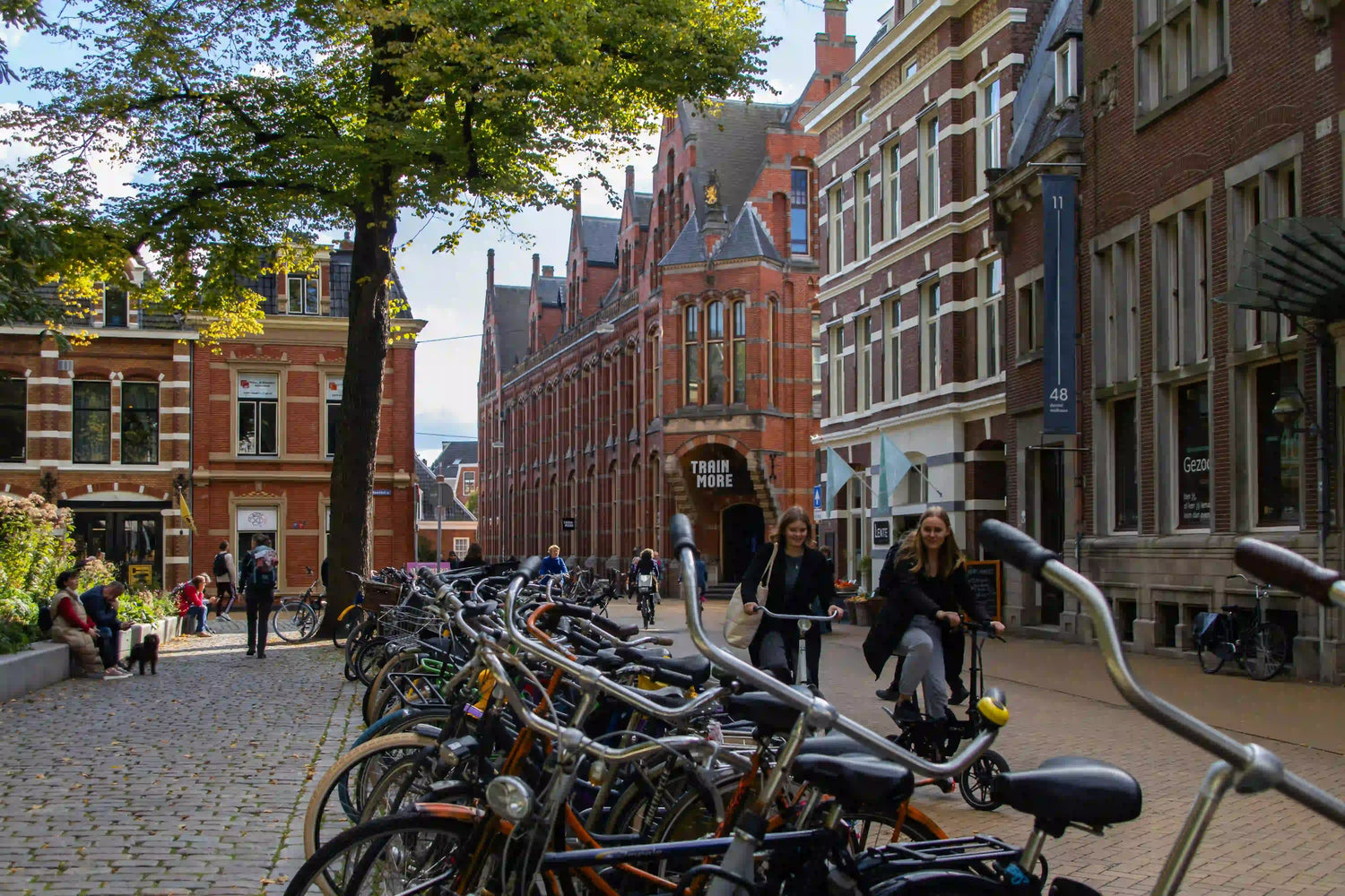 A bustling Dutch city square in Utrecht, with long rows of parked bicycles in the foreground and people, including two young women on an ebike, enjoying the lively, bike-friendly urban environment.