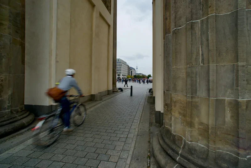 A cyclist in motion blur pedals an e-bike quickly through a narrow cobblestone archway in a European city