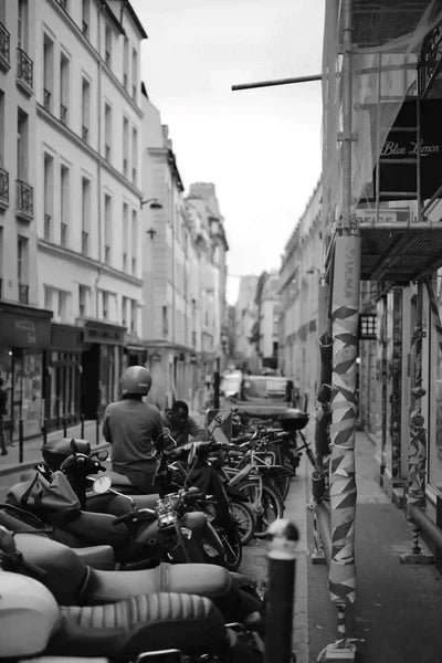 Black-and-white Paris side street with parked scooters and bicycles, a helmeted rider paused by the racks—showing the dense mix of two-wheelers in urban traffic 
