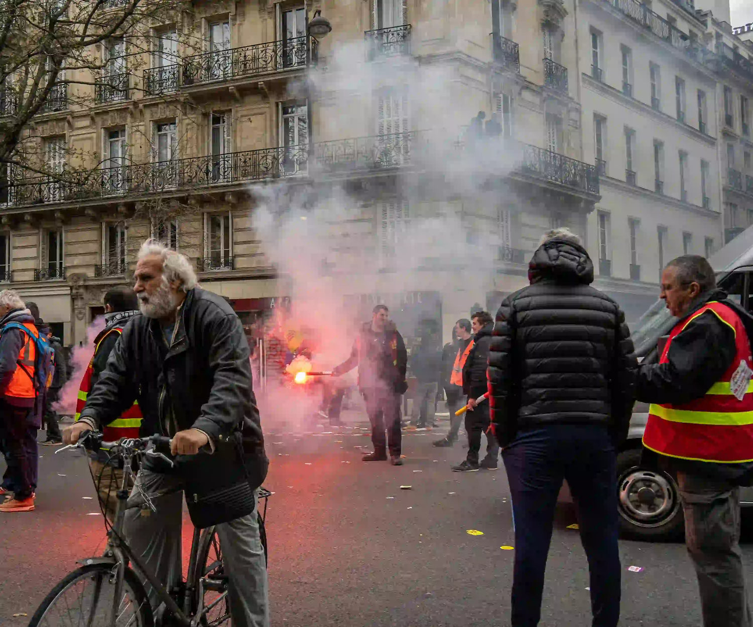 French street protest with smoke and flares, while a cyclist pushes his bike through the disruption.