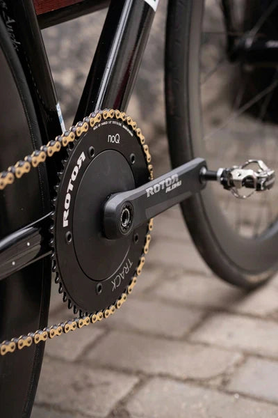 Close-up of a black bike crank and gold chain installed on a sleek matte black bicycle, showcasing a high-performance fixed gear component ideal for urban cycling and track racing.