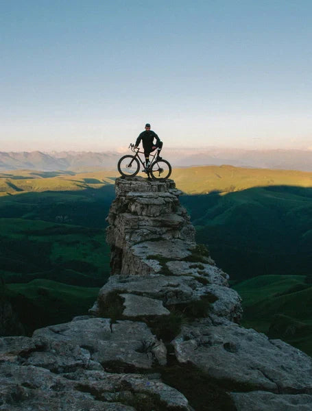 Cyclist standing with a gravel ebike on a rocky cliff summit overlooking a vast green landscape at sunset.